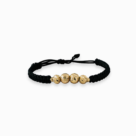 Pulsera tejida de color oscuro para adulto con cuatro balines de oro 18k de acabado diamantado (7mm y 6mm) sobre fondo blanco.
